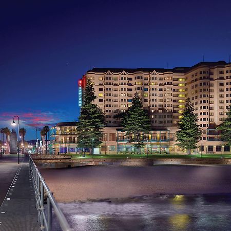Hotel Stamford Grand Adelaide Glenelg Esterno foto The photo shows a waterfront scene at dusk, featuring a large building lit up against a darkening sky. The architecture appears modern and prominent, with several windows illuminated. In the foreground, there is a walkway or pier with fencing along t