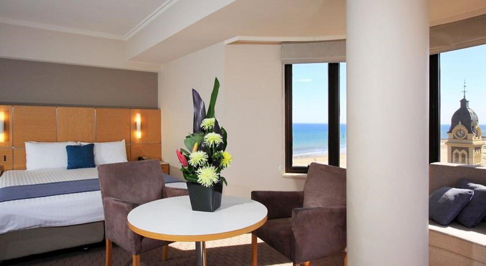 Hotel Stamford Grand Adelaide Glenelg Esterno foto The photo shows a cozy hotel room with a comfortable seating arrangement. There are two brown chairs positioned around a round table, which features a decorative flower arrangement in a black vase. The background includes a window that offers a view 