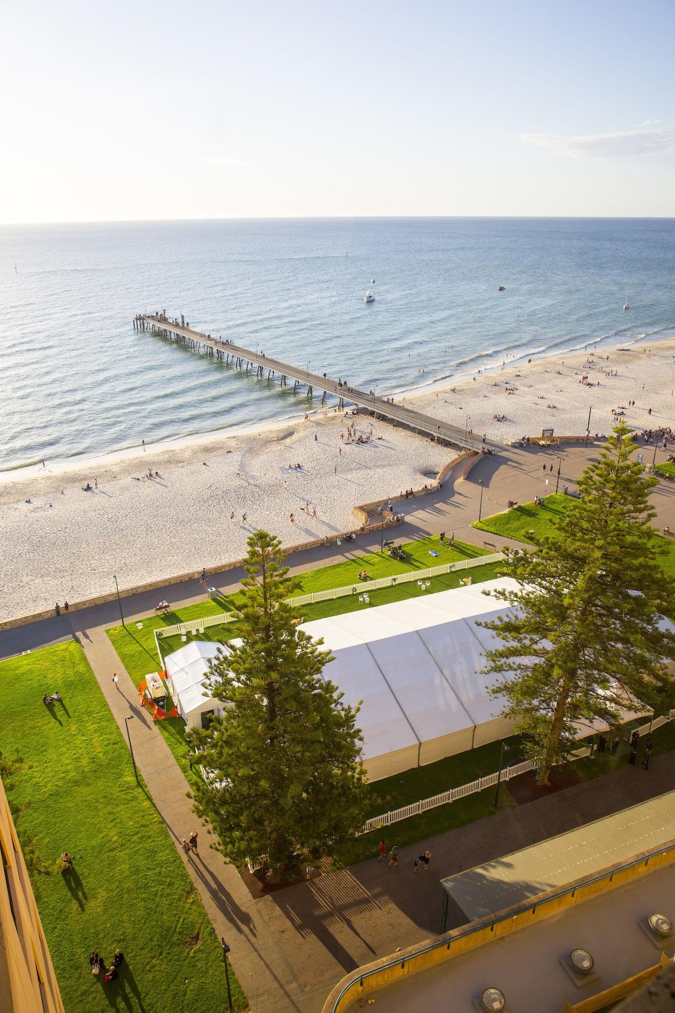 Hotel Stamford Grand Adelaide Glenelg Esterno foto The photo shows a scenic view of a beach with a long pier extending into the water. The beach is sandy, and there are people enjoying the sun and walking along the shore. In the foreground, there is a large white tent set up on a grassy area, likely 