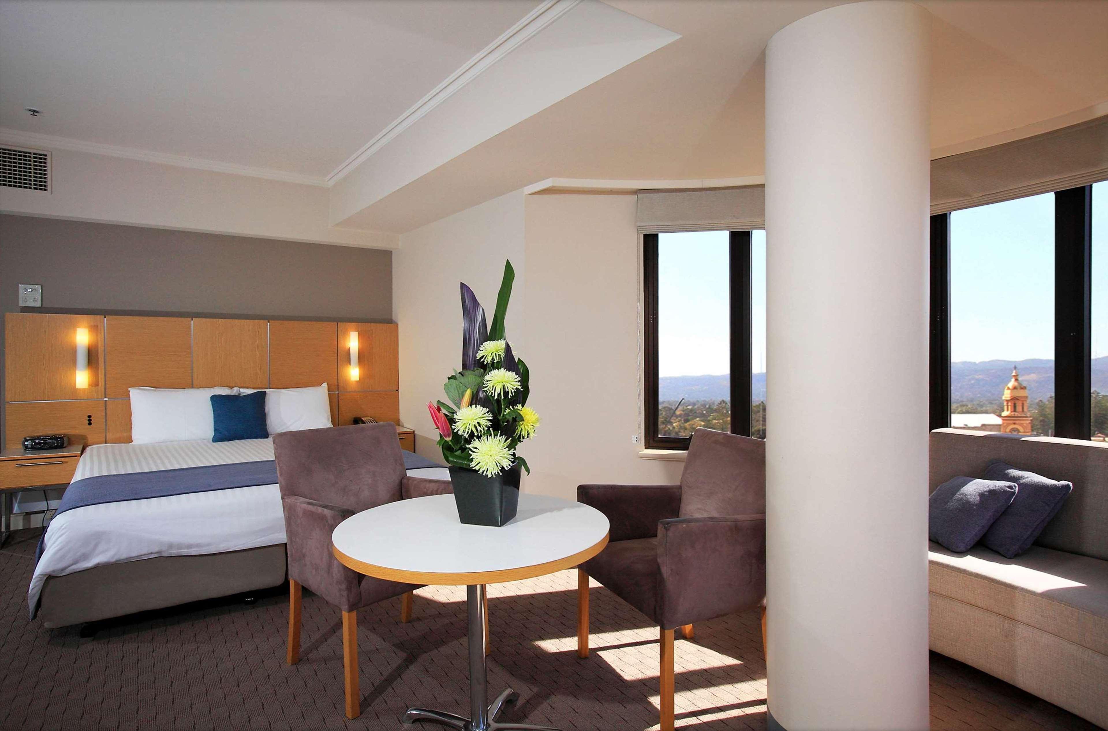 Hotel Stamford Grand Adelaide Glenelg Esterno foto The photo shows a hotel room with a modern design. On one side, there is a plush bed with white and blue bedding. In the center of the room, there's a small round table with two gray upholstered chairs around it. A vase of flowers sits on the table, 