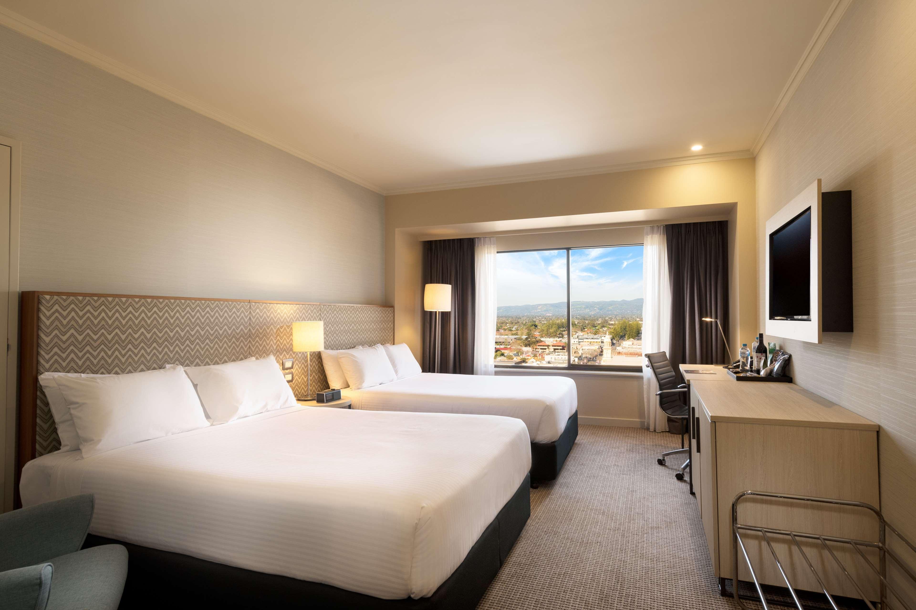 Hotel Stamford Grand Adelaide Glenelg Esterno foto The photo shows a modern hotel room featuring two beds with white linens. There are bedside tables with lamps next to each bed. A large window provides natural light and offers a view of the outside, with curtains on either side. The room has a neutr