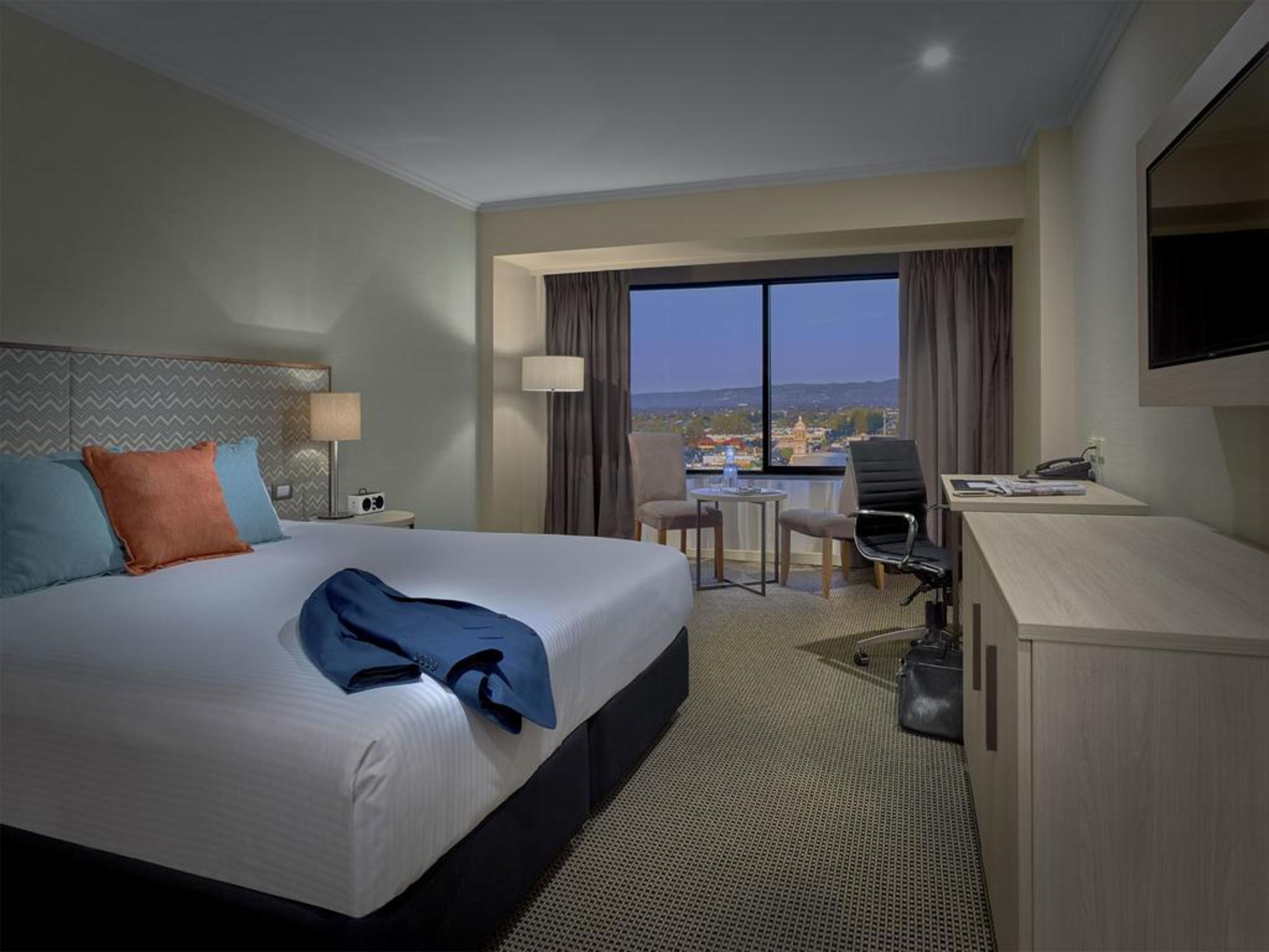 Hotel Stamford Grand Adelaide Glenelg Esterno foto The photo shows a modern hotel room. It features a large double bed with white bedding and decorative pillows in orange and blue. There's a nightstand with lamps on either side of the bed. A large window overlooks a view outside, and there's a desk a