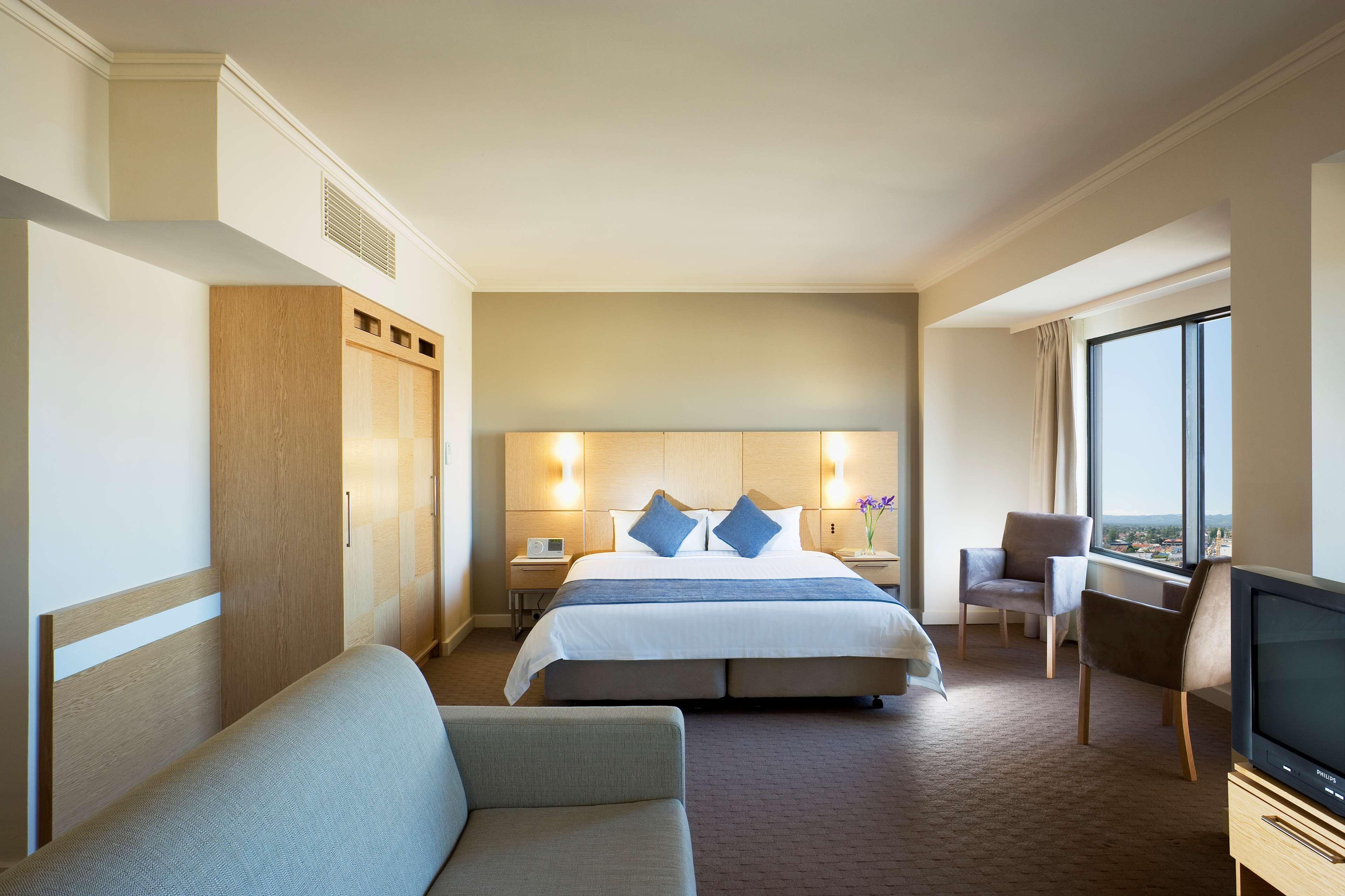 Hotel Stamford Grand Adelaide Glenelg Camera foto The photo shows a modern hotel room featuring a large bed with two blue decorative pillows. There are bedside lamps on both sides of the headboard. To the left, there's a wooden wardrobe and a small table with a vase and a few amenities. On the right