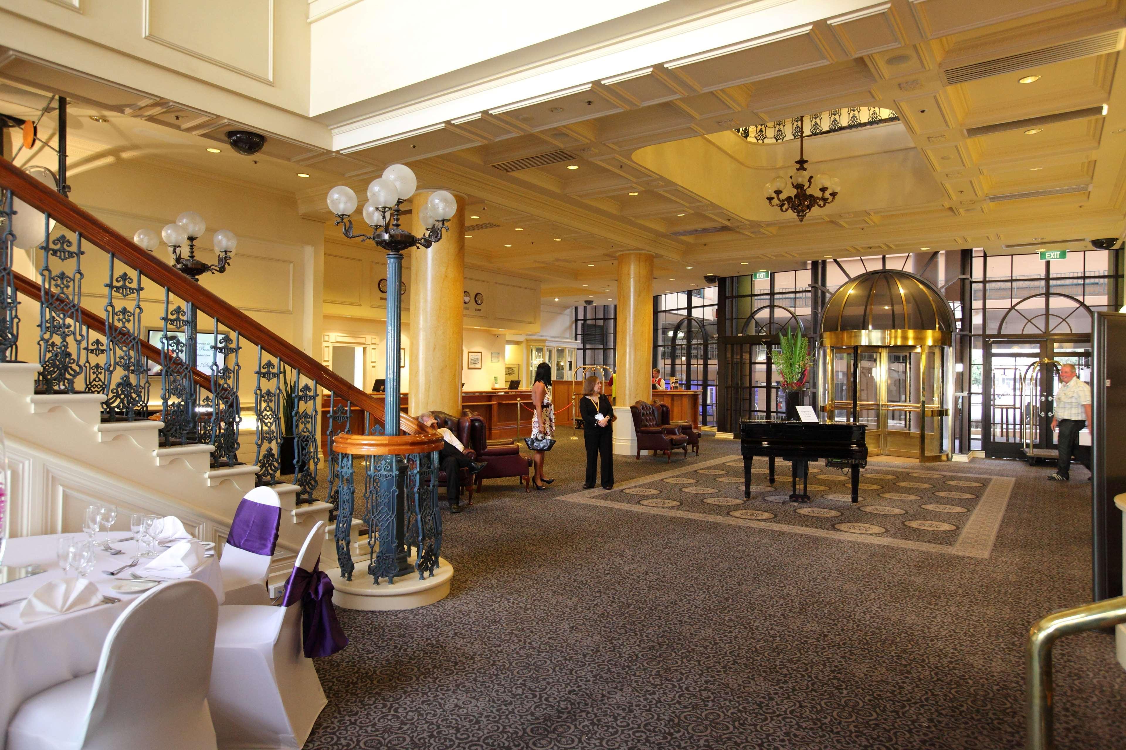 Hotel Stamford Grand Adelaide Glenelg Interno foto The photo shows a luxurious interior of a hotel or a restaurant lobby. There is an elegant staircase with ornate railings leading to an upper level. The space features high ceilings with decorative chandeliers and ample natural light coming through l