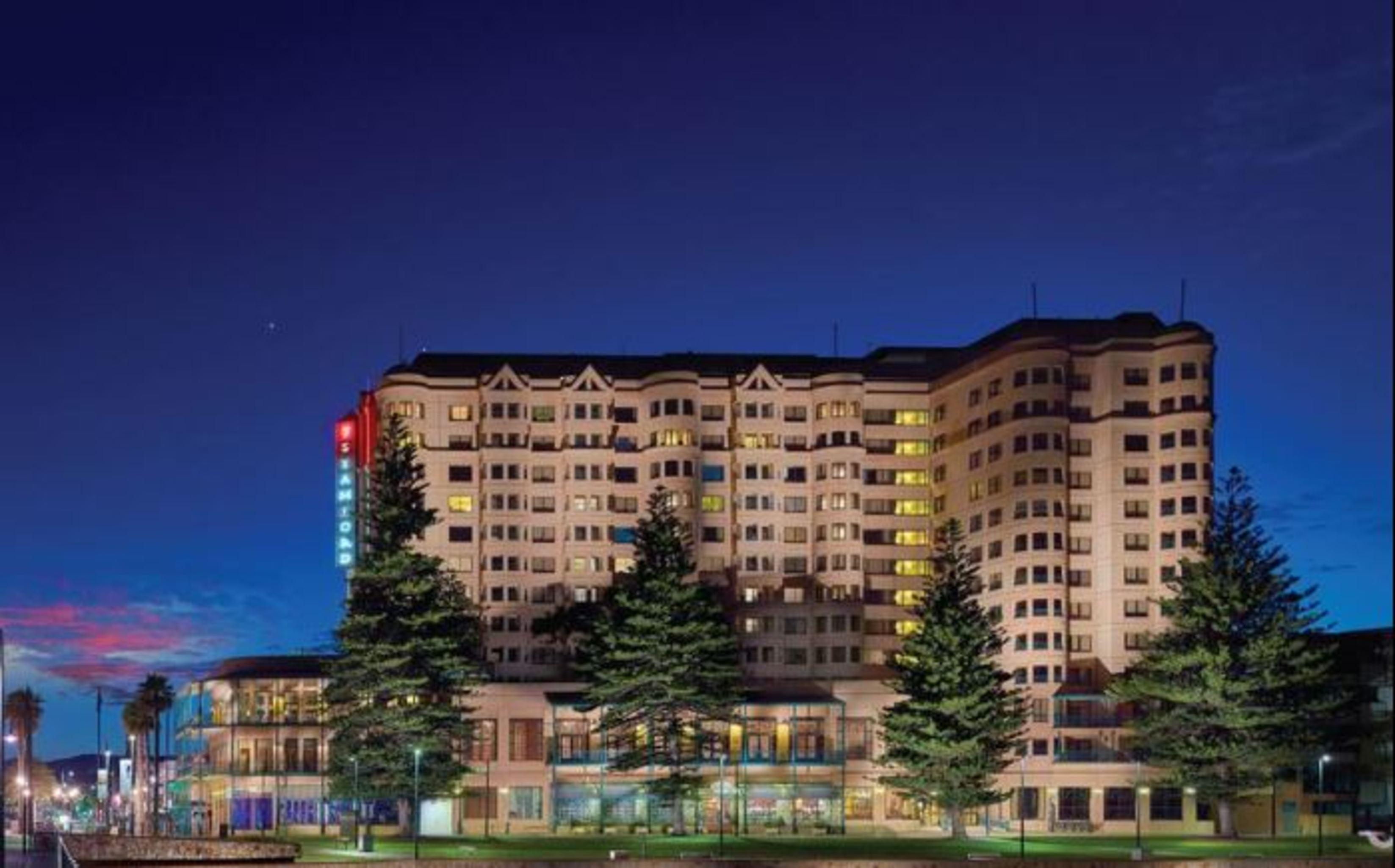 Hotel Stamford Grand Adelaide Glenelg Esterno foto The photo displays a multi-story hotel building at dusk, illuminated by various lights. The architecture features a mix of modern and traditional design elements, with numerous windows showcasing a warm glow. Surrounding the hotel are several tall tr