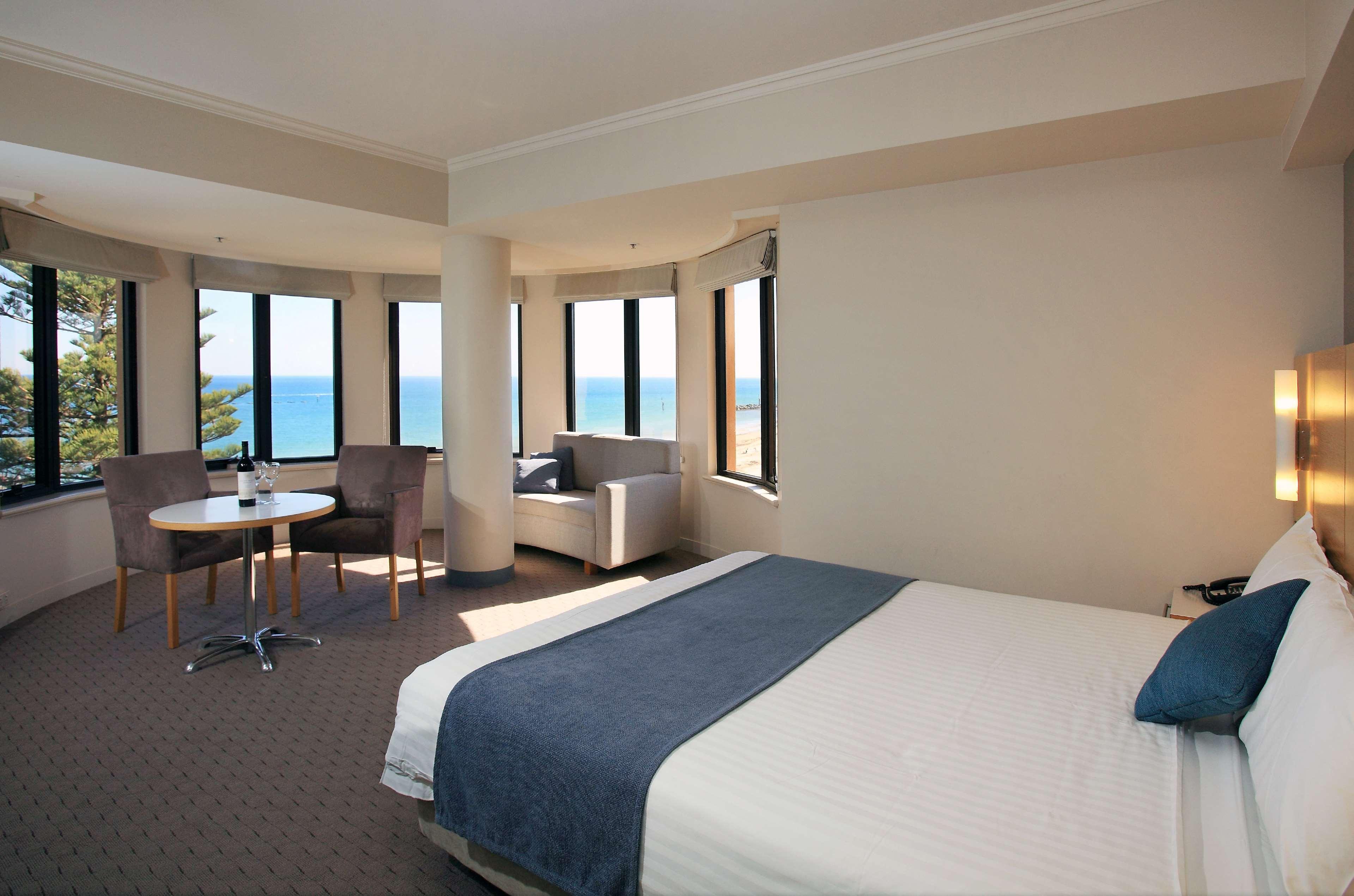 Hotel Stamford Grand Adelaide Glenelg Esterno foto The photo shows a well-lit hotel room featuring large windows that offer a view of the ocean. The room is arranged with a comfortable bed covered in white linens and a navy blue throw blanket. There is a small table and chairs set up beside the windo