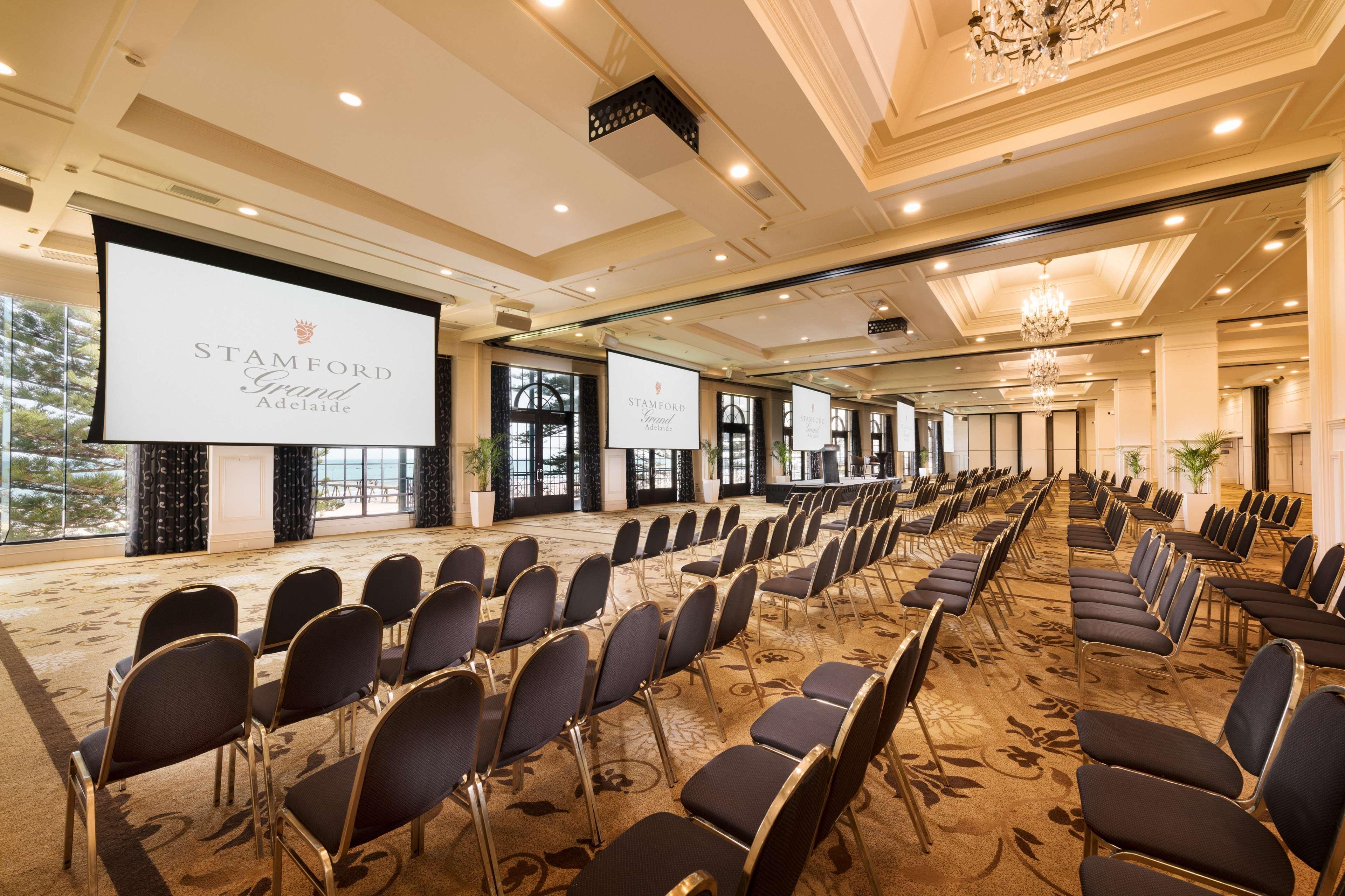 Hotel Stamford Grand Adelaide Glenelg Esterno foto The photo shows a spacious event or conference room. There are rows of dark, upholstered chairs arranged facing two large screens. The room features elegant decor with soft lighting, a chandelier, and windows that allow natural light to filter in. Th