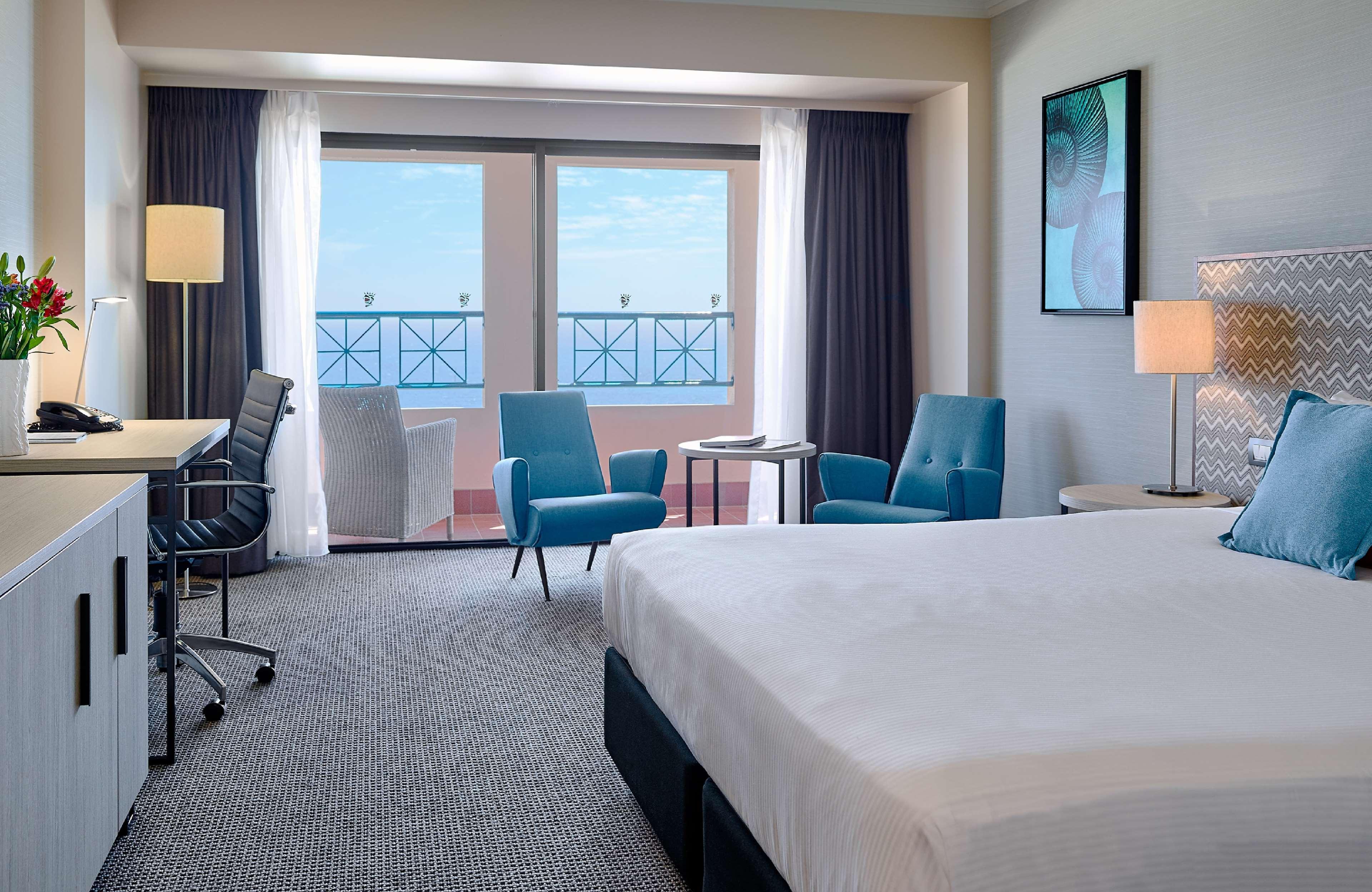 Hotel Stamford Grand Adelaide Glenelg Esterno foto The image shows a hotel room featuring a large bed with a white duvet. There are two bright blue chairs and a small table near a large window that provides a view outside, possibly to a balcony. The room includes a workspace with a desk and an office