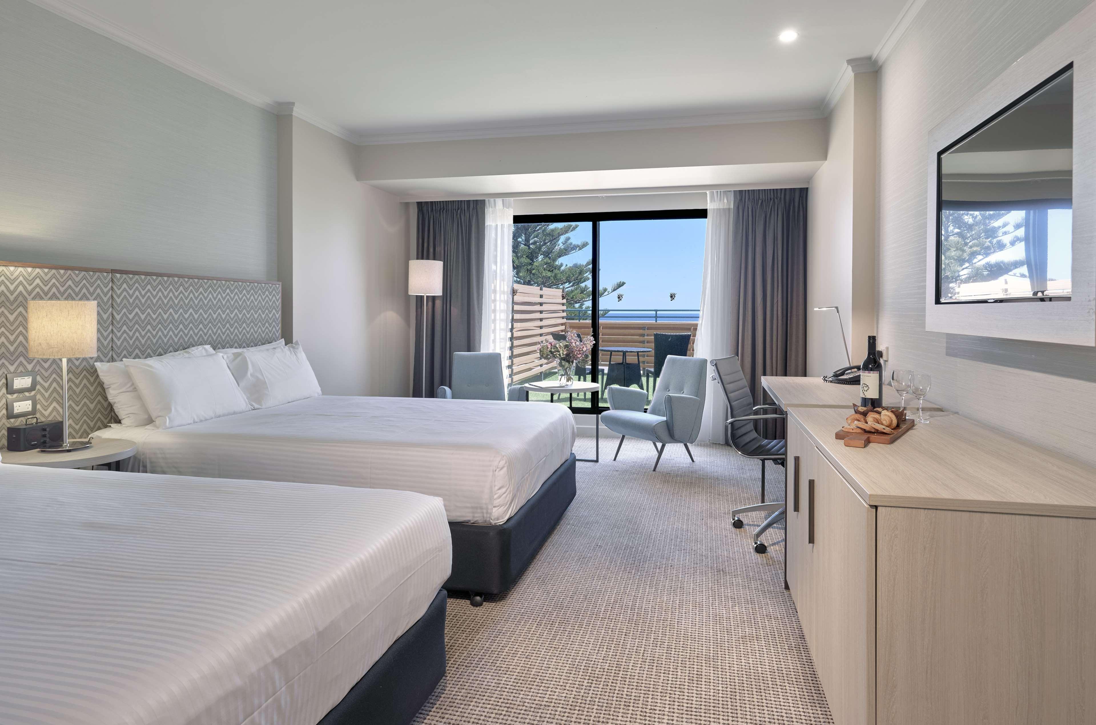 Hotel Stamford Grand Adelaide Glenelg Esterno foto The photo shows a modern hotel room featuring two beds with white linens. There's a large window that provides natural light and a view of a balcony area. The room includes two chairs and a small table, creating a cozy sitting area. A television is m